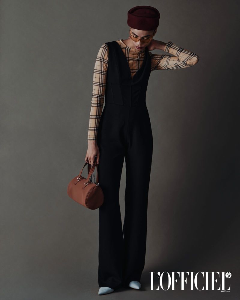 Model Vitorinni poses in a black sleeveless jumpsuit over a plaid top, wearing a maroon pillbox hat against a grey seamless backdrop.