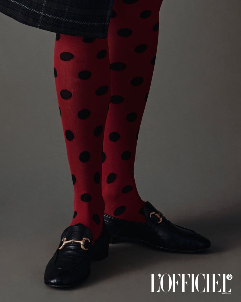 Close-up of legs and feet wearing red polka dot tights and black leather loafers with gold hardware against a grey backdrop.