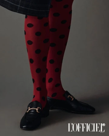 Close-up of legs and feet wearing red polka dot tights and black leather loafers with gold hardware against a grey backdrop.