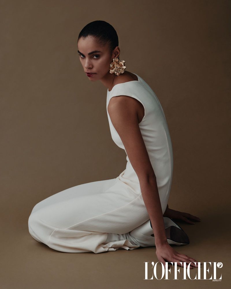Fashion model in a white jumpsuit and large gold earrings poses kneeling in a studio with tan seamless background looking at camera.