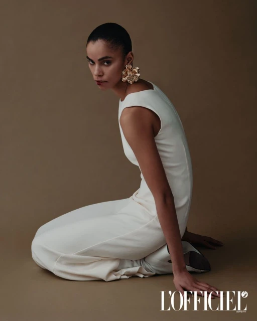 Fashion model in a white jumpsuit and large gold earrings poses kneeling in a studio with tan seamless background looking at camera.