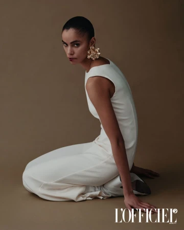 Fashion model in a white jumpsuit and large gold earrings poses kneeling in a studio with tan seamless background looking at camera.
