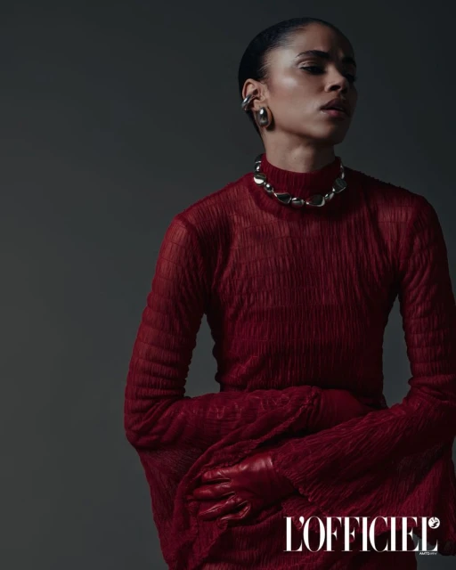 Fashion editorial shot of a young adult woman wearing a sheer red top, gloves, and silver jewelry against a dark gray background.