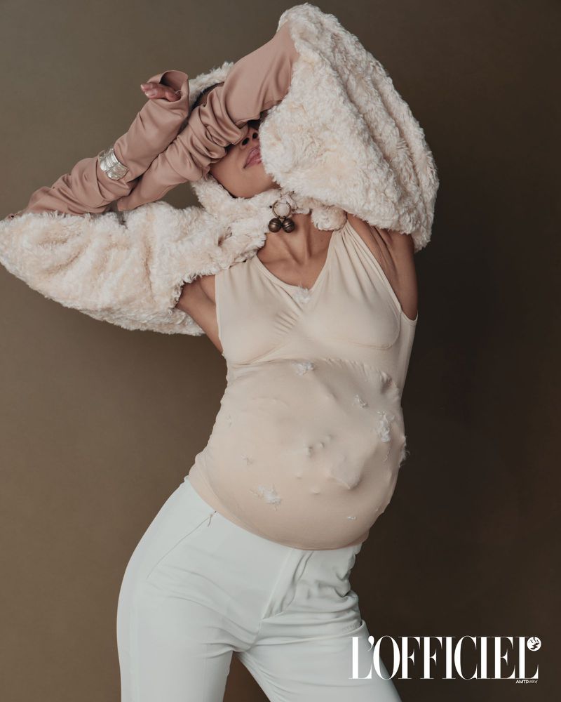 Editorial fashion shot of a posing adult female wearing a cream colored bodysuit with white pants against a brown background.