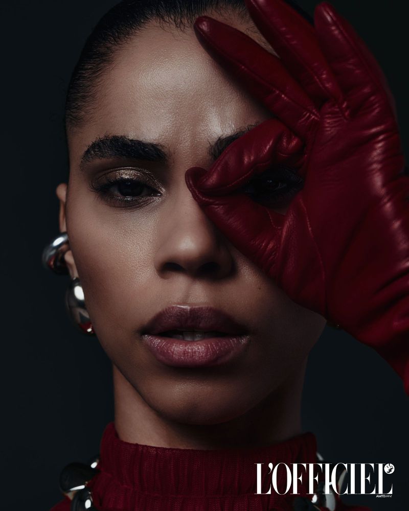 Close-up studio shot of a dark-skinned adult woman with red leather glove held to her face, statement jewelry, and dark background.