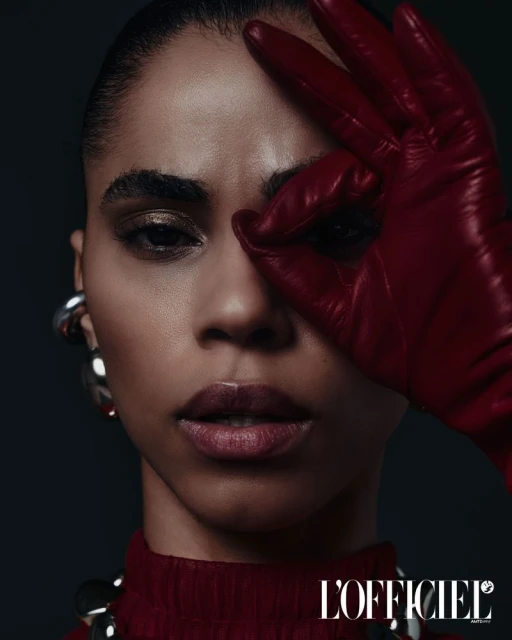 Close-up studio shot of a dark-skinned adult woman with red leather glove held to her face, statement jewelry, and dark background.