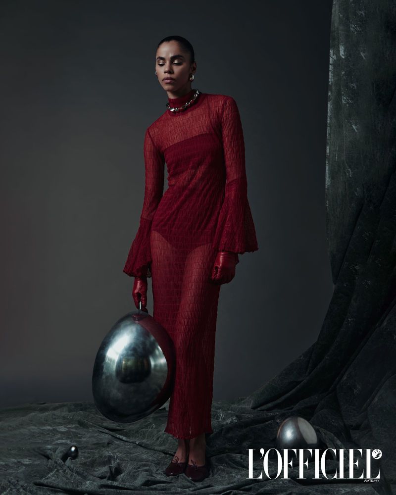 Fashion editorial shot of a slender adult female model in a red sheer dress with gloves, holding a metallic silver orb in studio.