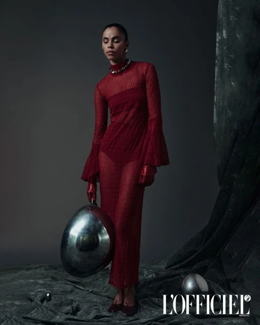 Fashion editorial shot of a slender adult female model in a red sheer dress with gloves, holding a metallic silver orb in studio.