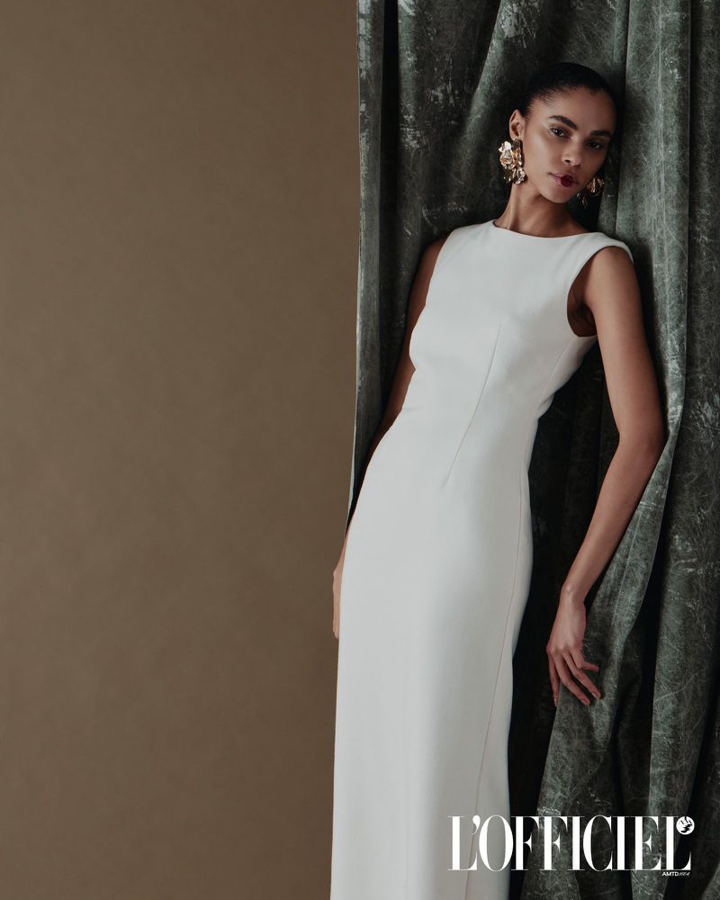Elegant fashion shot of an adult woman wearing a white sleeveless gown, leaning against a green curtain in a neutral indoor setting.