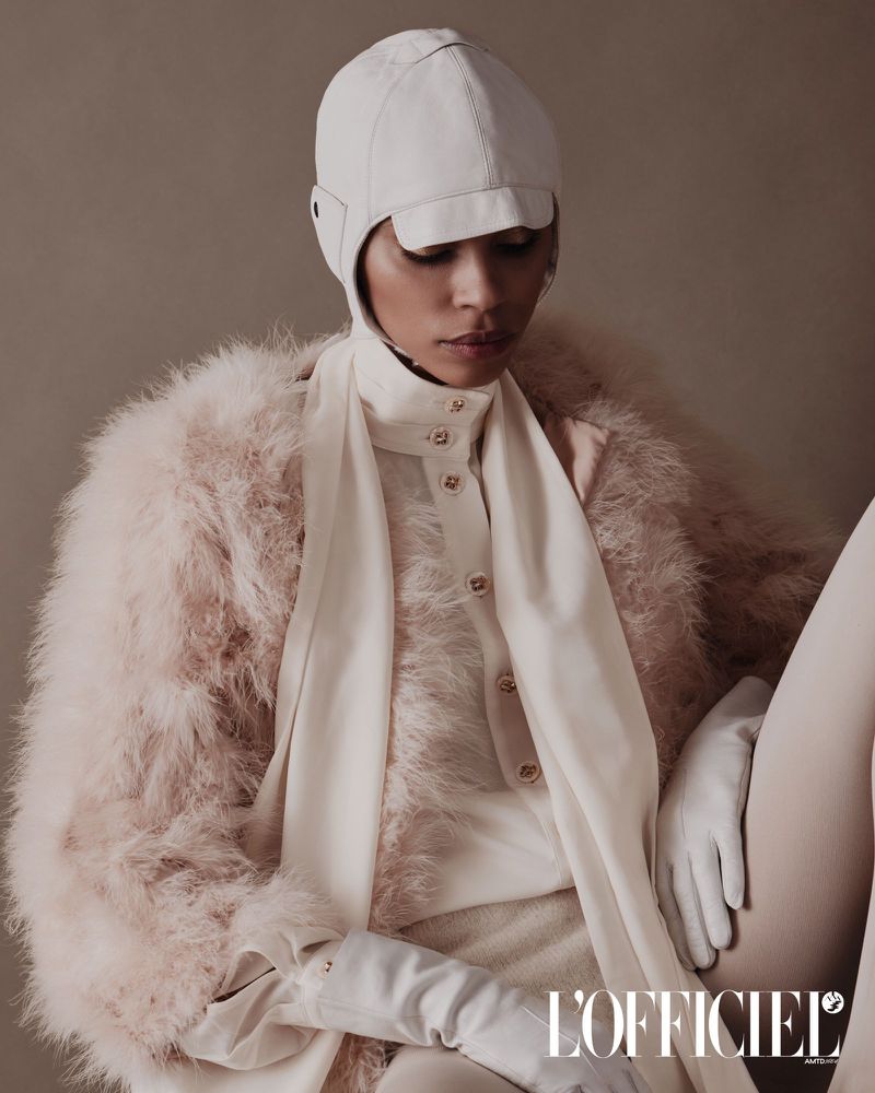 Model Vitorinni seated wearing a pale fuzzy feathered coat, white bonnet cap, and white gloves against a neutral background.