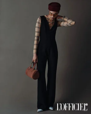 Model Vitorinni poses in a black sleeveless jumpsuit over a plaid top, wearing a maroon pillbox hat against a grey seamless backdrop.