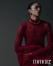 Model Vitorinni wearing a textured burgundy long-sleeve dress, red leather gloves, and silver jewelry against a dark grey backdrop.