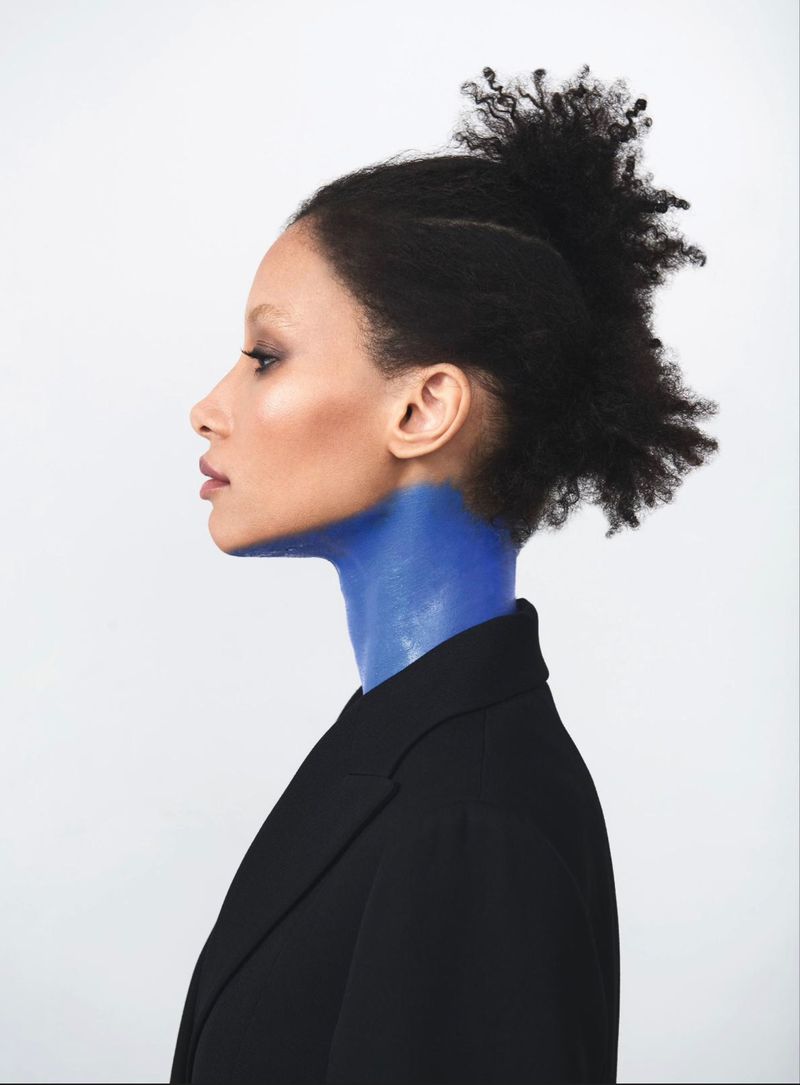 Profile of an adult woman with dark hair wearing a black blazer, her neck and upper chest painted blue against a white background.