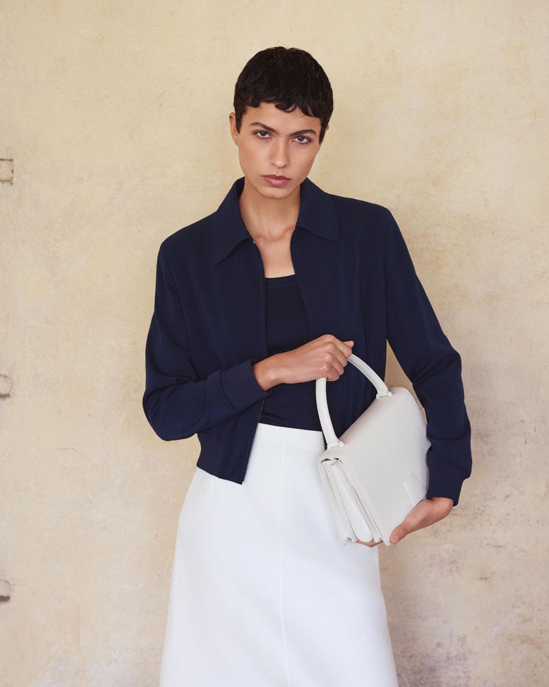 Woman in navy jacket and white skirt holds a white handbag against a textured neutral background in a fashion studio shot.