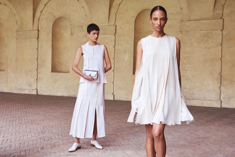 Two women in sleeveless white dresses stand in an arched hallway. One holds a clutch. The scene is lit by natural light.