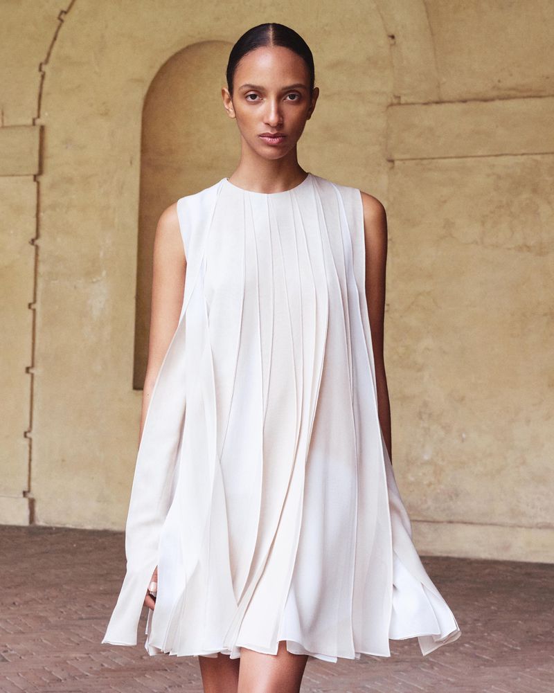 A slender adult woman is posing in a sleeveless pleated dress against a light, neutral-colored, arched architectural backdrop with soft, diffused light.