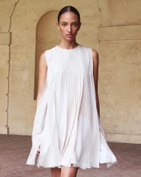 Model wearing a sleeveless white pleated mini dress standing in front of a warm stone archway.