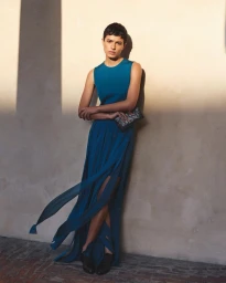 Model with short pixie hair wearing a sleeveless teal dress with flowing slit skirt panels against a sunlit wall.