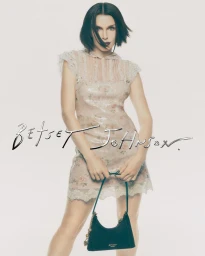 Julia Fox wearing a floral minidress and holding a black handbag against a white studio background for the Betsey Johnson campaign.