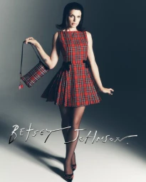 Julia Fox in a red plaid minidress holding a matching handbag against a grey studio background for Betsey Johnson Holiday 2026 campaign.
