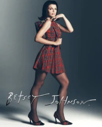 Julia Fox wearing a red plaid dress and matching heels holding a handbag against a grey backdrop for Betsey Johnson Holiday 2026