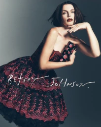 Julia Fox in a black and red lace dress holding a clutch against a grey seamless background for Betsey Johnson Holiday 2026