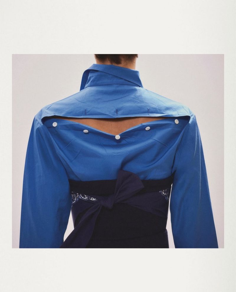 Back view of a person in a blue button-down shirt with an open back and a black obi sash against a neutral backdrop.