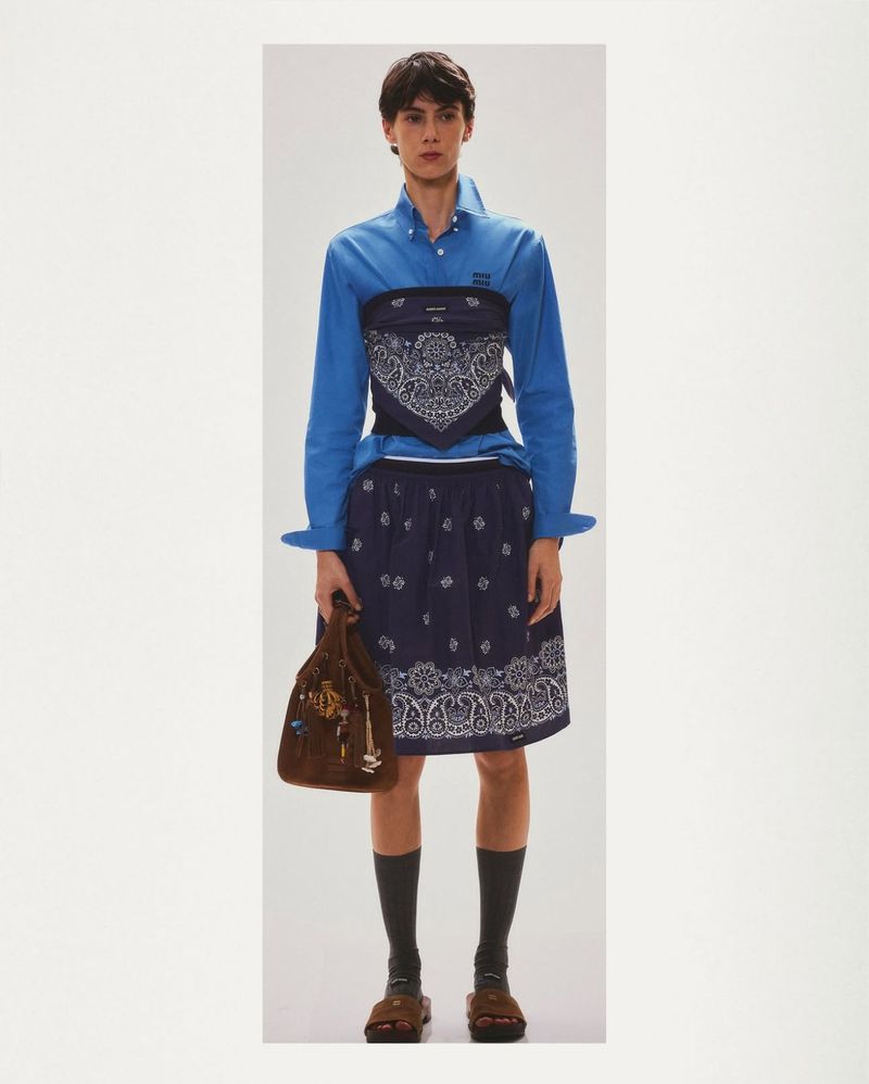 Full-length studio shot: a person in a blue shirt, blue bandana skirt, and brown sandals stands against a seamless white backdrop.