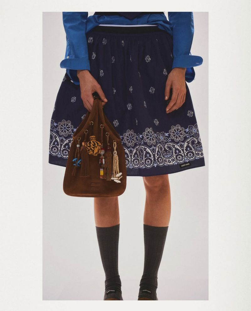 A person holds a brown purse, wearing a blue shirt, dark blue skirt with white paisley, and dark socks, against a neutral background.