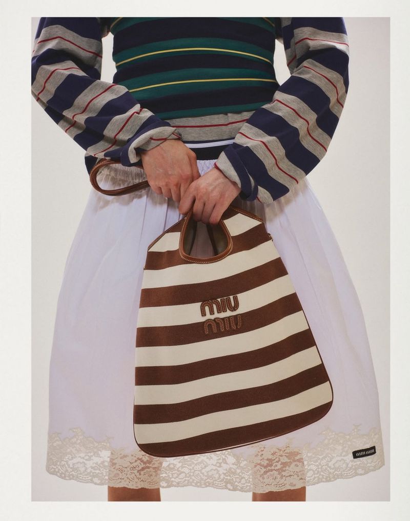 Striped Miu Miu tote bag held by a person wearing a striped long-sleeve shirt and white skirt with lace trim, studio shot.