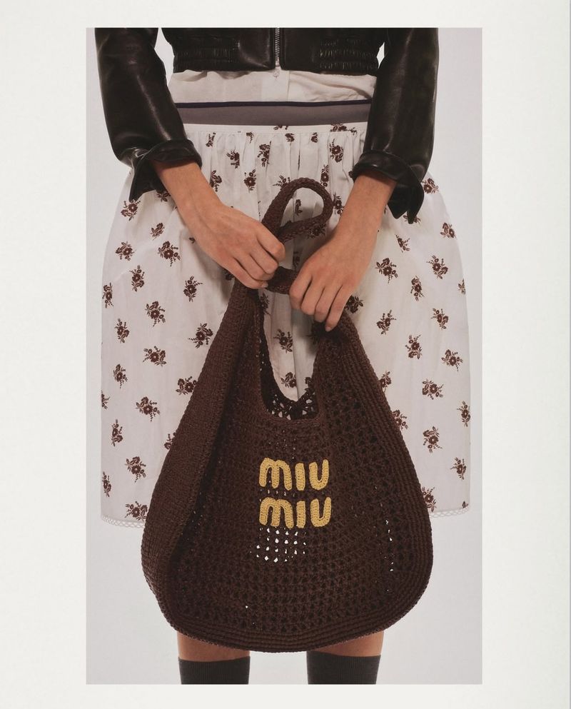 A model showcases a brown crocheted Miu Miu handbag against a white backdrop, accessorized with a black leather jacket and patterned skirt.