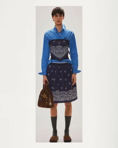 Model posing in a blue button-down shirt layered with a navy bandana-print corset and matching skirt against a studio backdrop.