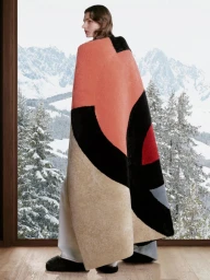 Model standing indoors wrapped in a large geometric color-blocked blanket with a snowy mountain backdrop visible through a window.