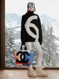 Model wearing a black sweater with white logo, white pants, and holding a colourful quilted bag against a snowy mountain view.