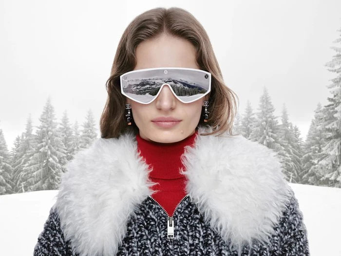 Model wearing white mirrored ski goggles, red turtleneck, and tweed jacket with fur collar against a snowy forest backdrop