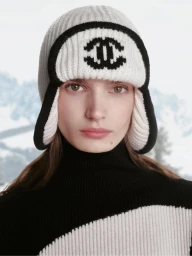 Model wearing a white and black knitted trapper hat with the Chanel logo and a black turtleneck sweater against a snowy mountain backdrop.
