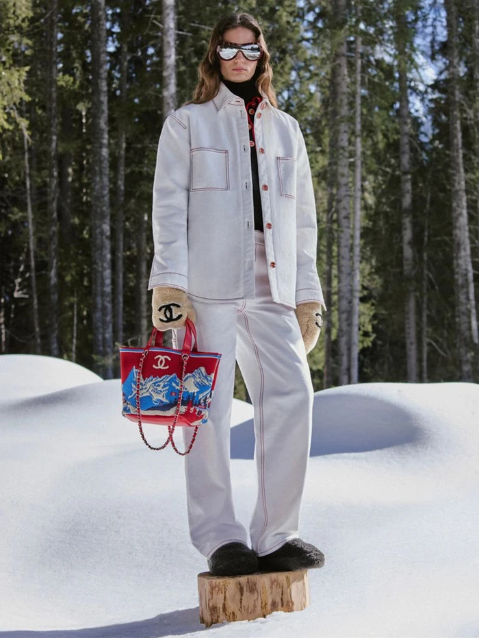 Model standing on a tree stump in a snowy forest wearing a white jacket, trousers, sunglasses, gloves, and holding a red handbag.