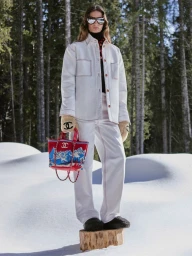 Model standing on a tree stump in a snowy forest wearing a white jacket, trousers, sunglasses, gloves, and holding a red handbag.
