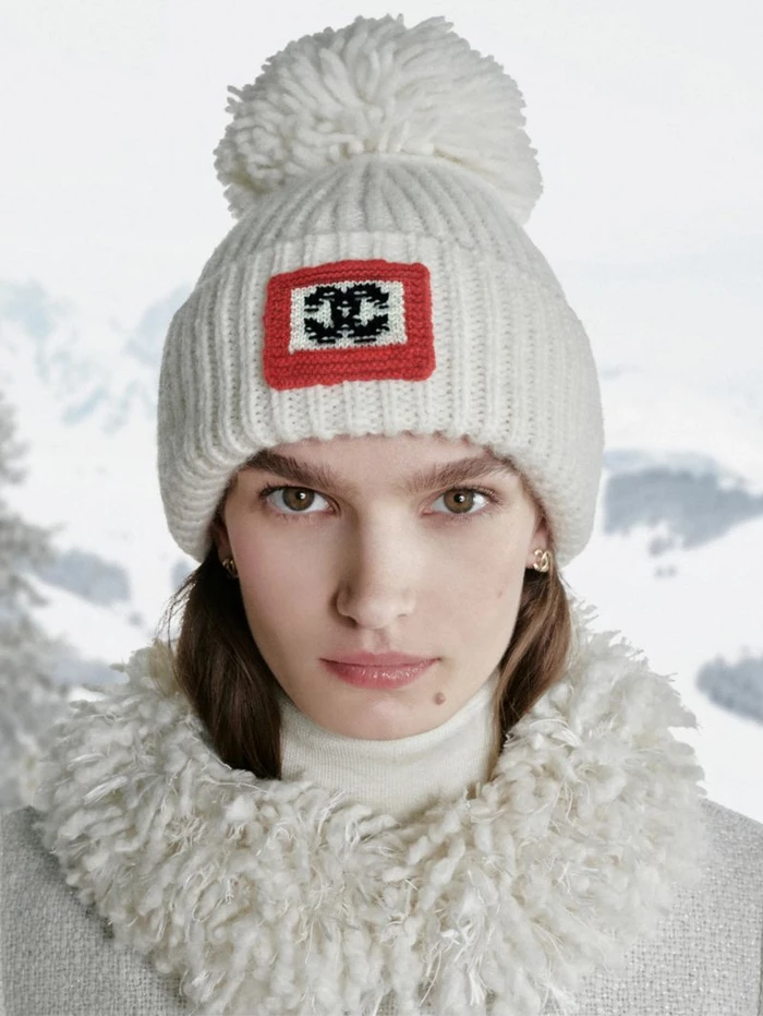 Model wearing a white knit beanie with a pom-pom and a CC logo patch, photographed outdoors against a snowy mountain landscape.