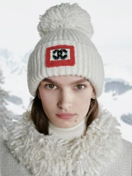 Model wearing a white knit beanie with a pom-pom and a CC logo patch, photographed outdoors against a snowy mountain landscape.
