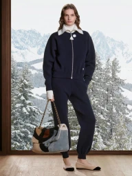 Model standing indoors before a snowy mountain window holding a shearling chain handbag wearing a navy zip-up tracksuit.