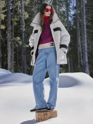 Model standing on a wooden stump in a snowy forest wearing a white jacket, patterned sweater, and blue jeans