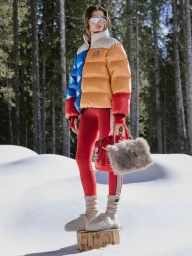 Model wearing a colour-blocked puffer jacket and red leggings standing on a tree stump in a snowy forest.