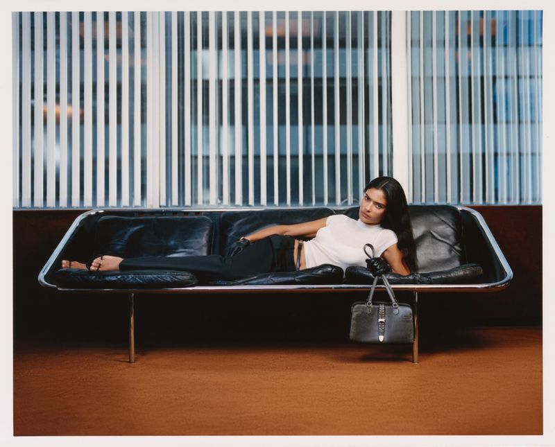Model lying on a black leather sofa holding a handbag in front of vertical blinds for Kith Women Autumn 2026