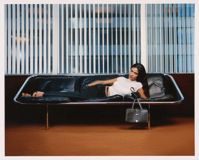 Model lying on a black leather sofa holding a handbag in front of vertical blinds for Kith Women Autumn 2026