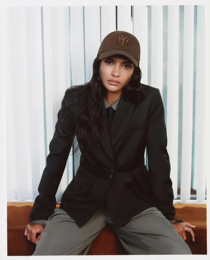 Model Rania Benchegra in a black blazer and brown baseball cap seated against white vertical blinds for Kith Women's Autumn 2026