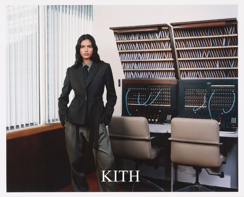 Model wearing a dark belted blazer and trousers standing in a vintage office interior with switchboards