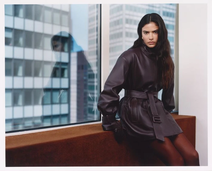 Model Rania Benchegra seated against a window with city reflection wearing a belted leather coat for Kith