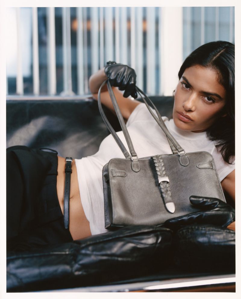 Model reclining on a dark leather couch holding a grey leather handbag for Kith Women