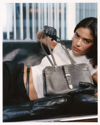 Model reclining on a dark leather couch holding a grey leather handbag for Kith Women